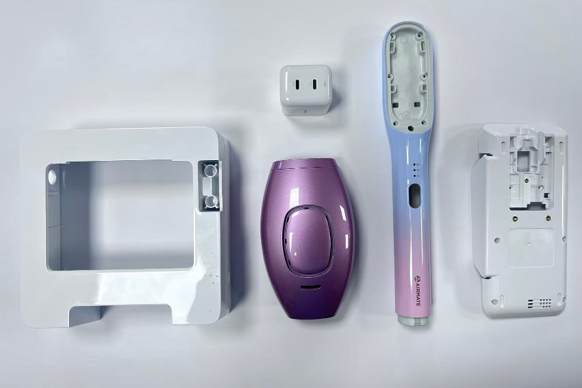 Six injection molded housing parts on an inspection bench at JBRplas — a white panel housing, a high-gloss purple device shell, a blue-purple gradient handle housing, and an inner frame with ribs and screw bosses